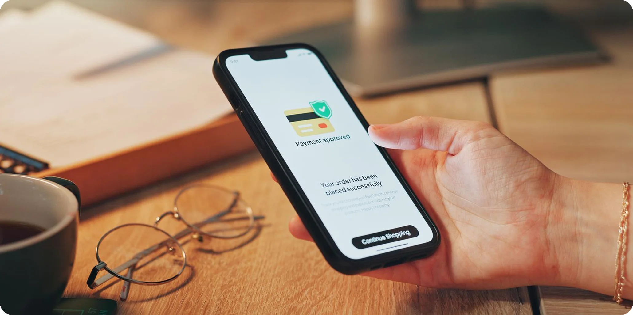Hand holding a smartphone displaying a payment approved confirmation screen with a credit card icon and a 'Continue Shopping' button.