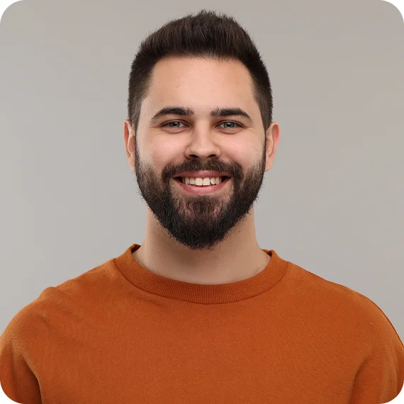 Smiling bearded man with short dark hair wearing an orange sweater against a plain gray background.