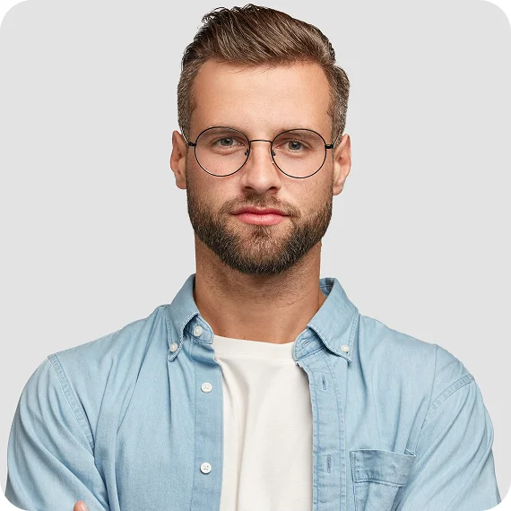 Young man with short brown hair, beard, and glasses wearing a light blue denim shirt over a white t-shirt against a gray background.