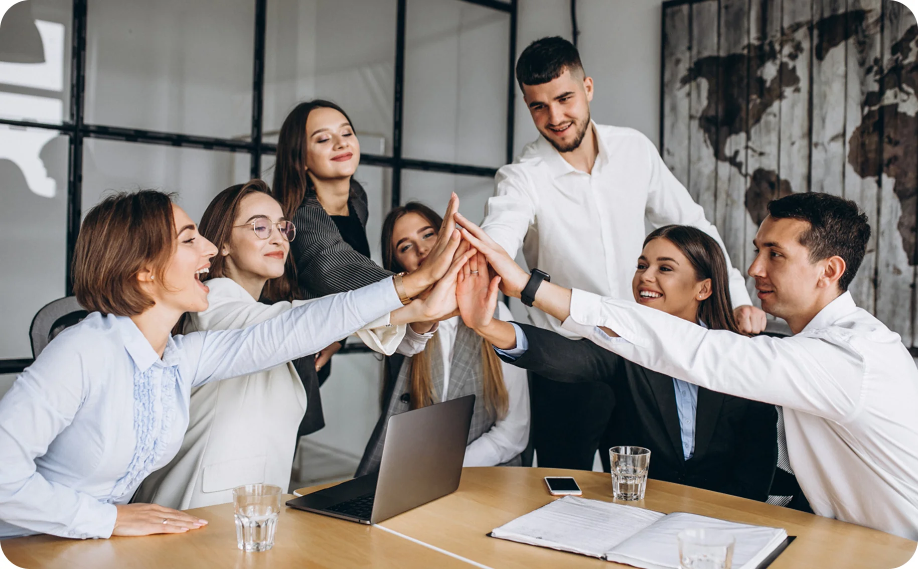 Group of six diverse young professionals in business attire giving a team high-five around a conference table with a laptop and documents.