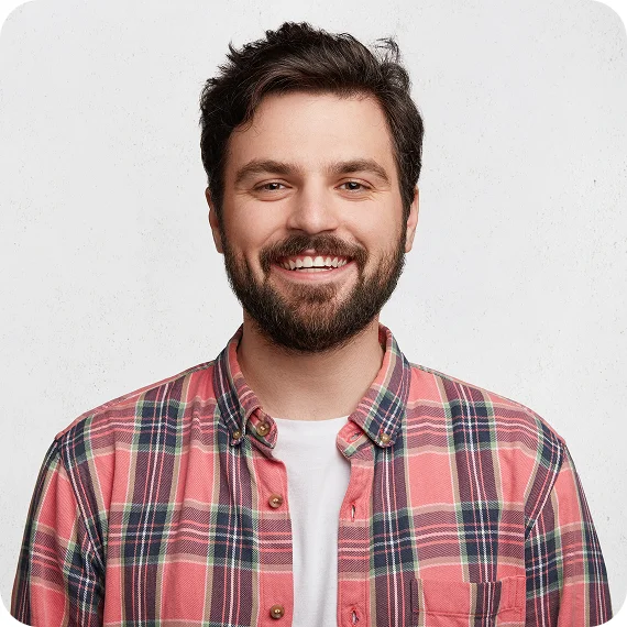Smiling man with dark hair and beard wearing a red and black plaid shirt over a white t-shirt against a light background.