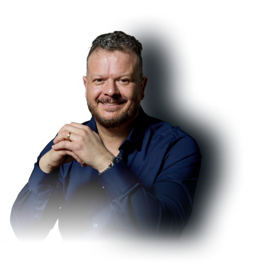Homem sorridente de camisa azul escura com barba e cabelo curto, com as mãos entrelaçadas.