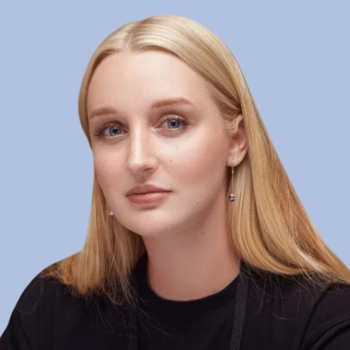 Portrait of a young woman with straight dark brown hair, wearing a black shirt, smiling against a light blue background.