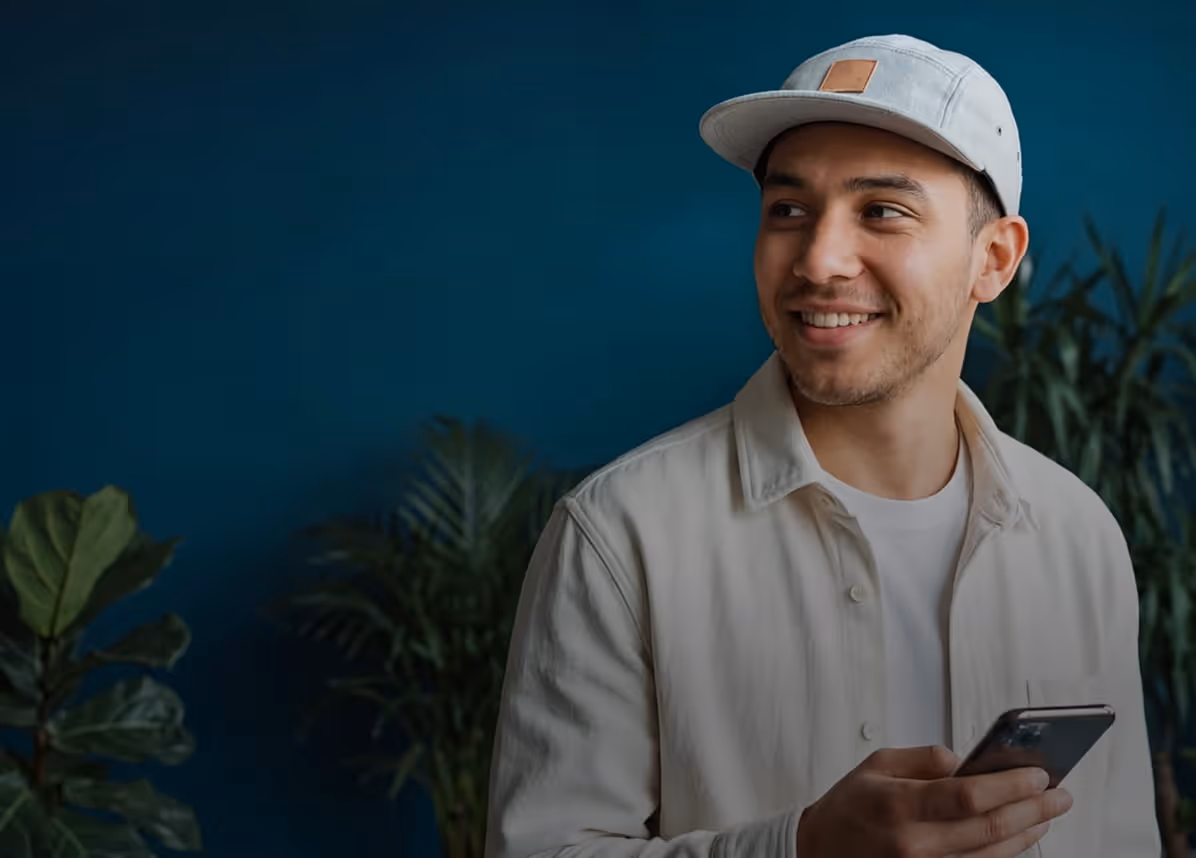 Young man in light jacket and cap smiling while holding a smartphone with plants in the background.