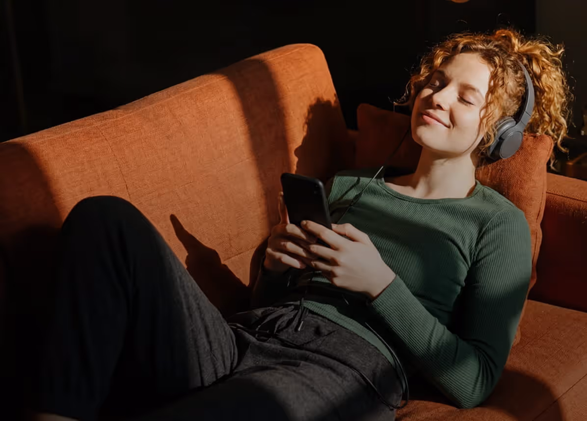 Young woman with curly hair relaxing on a rust-colored sofa, wearing headphones and holding a smartphone with eyes closed.
