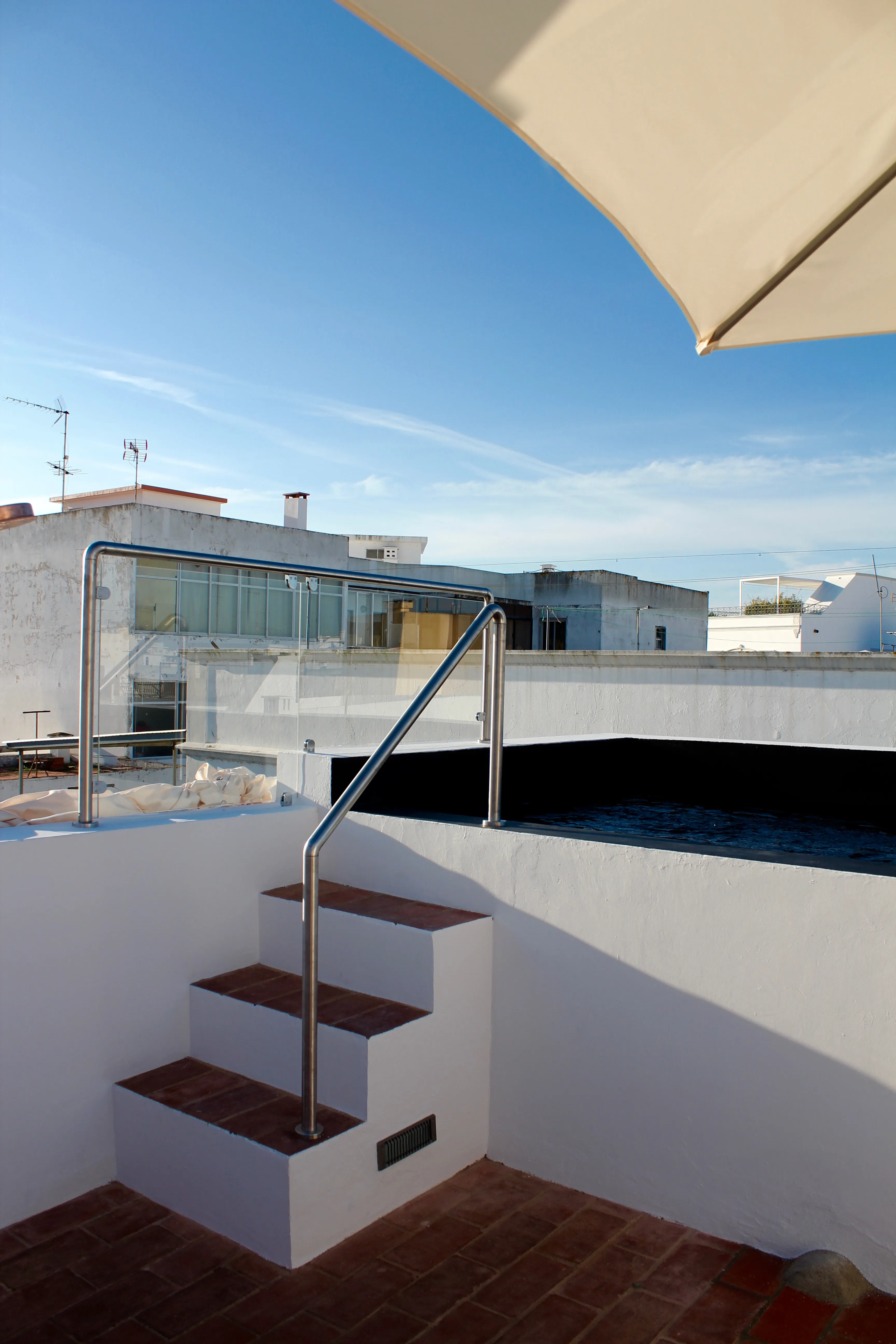 Small outdoor pool with white walls and tiled steps under a clear blue sky, adjacent to white buildings.