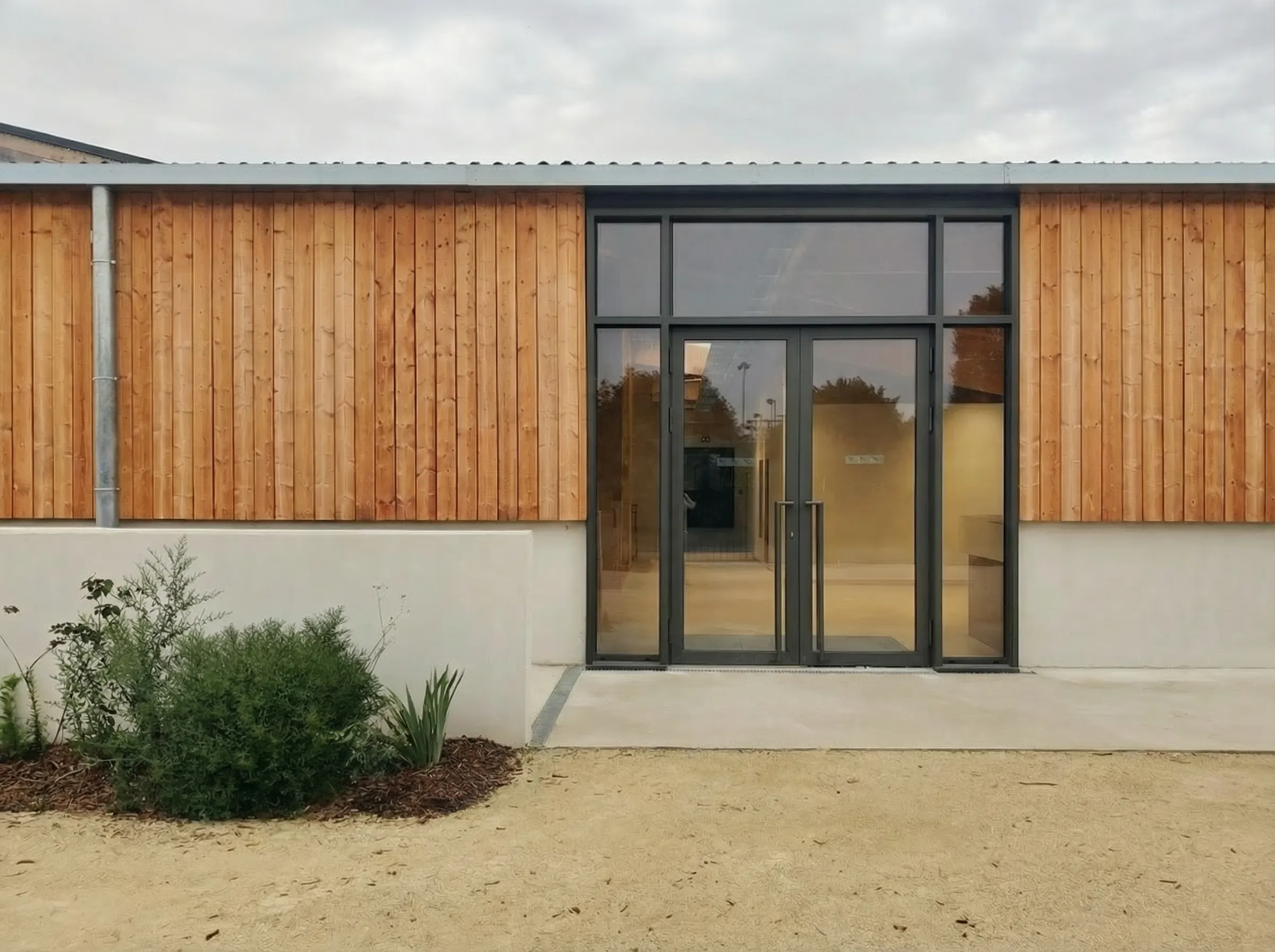 Entrée d'un bâtiment moderne avec un mur en bois clair, une porte vitrée noire et un parterre de plantes vertes devant.