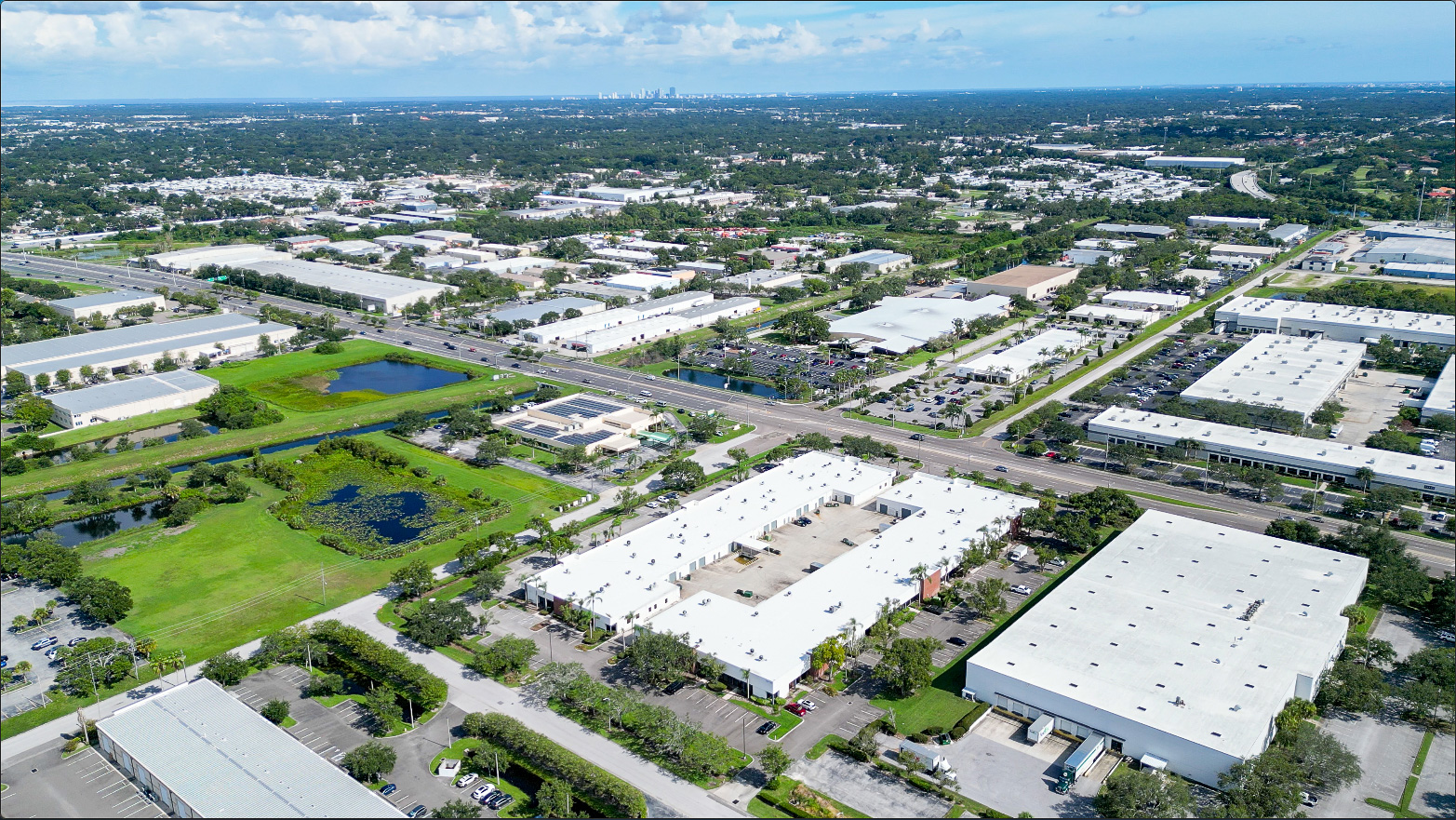 Aerial view of an industrial area with multiple white-roofed warehouses, roads, green spaces, and small water bodies under a partly cloudy sky.