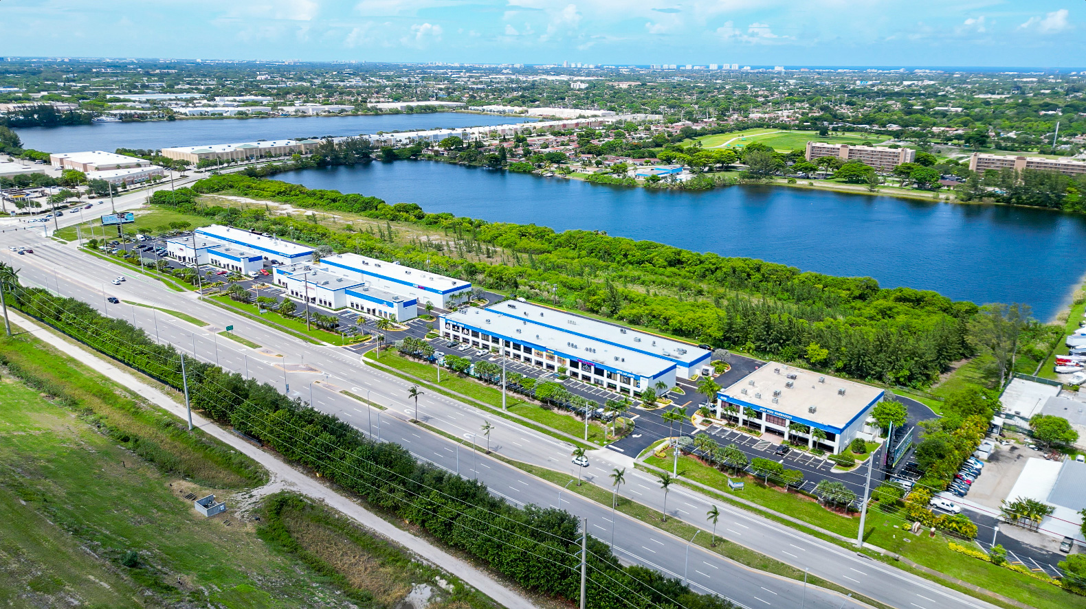 Aerial view of a business park with multiple rectangular white and blue-roofed buildings alongside a large road and a lake surrounded by greenery.