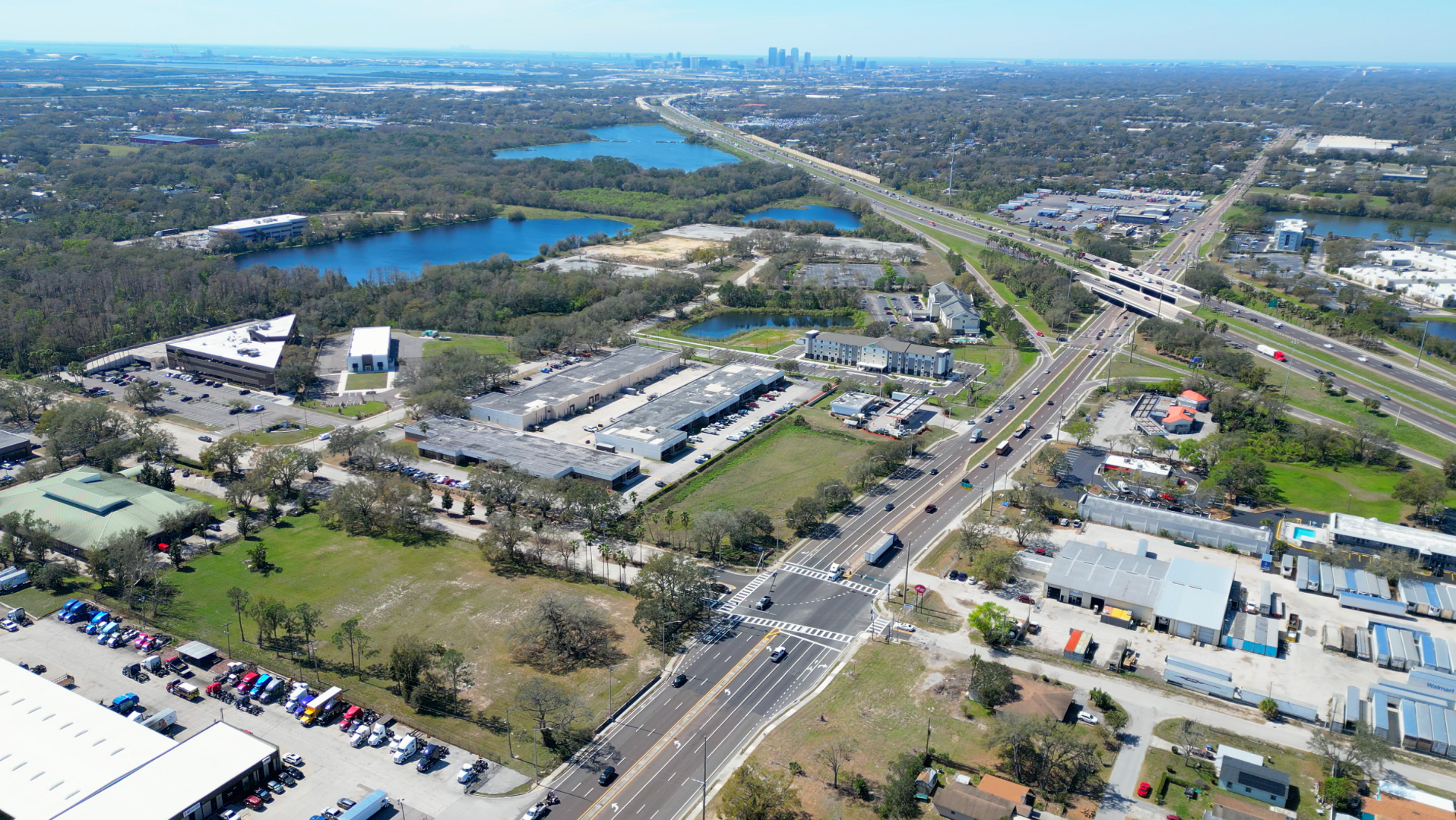 Aerial view of an urban area with a highway intersection, commercial buildings, lakes, and green spaces under a clear blue sky.