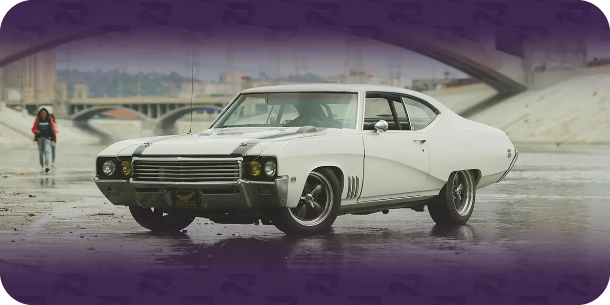 White classic muscle car parked on wet pavement in front of a concrete bridge