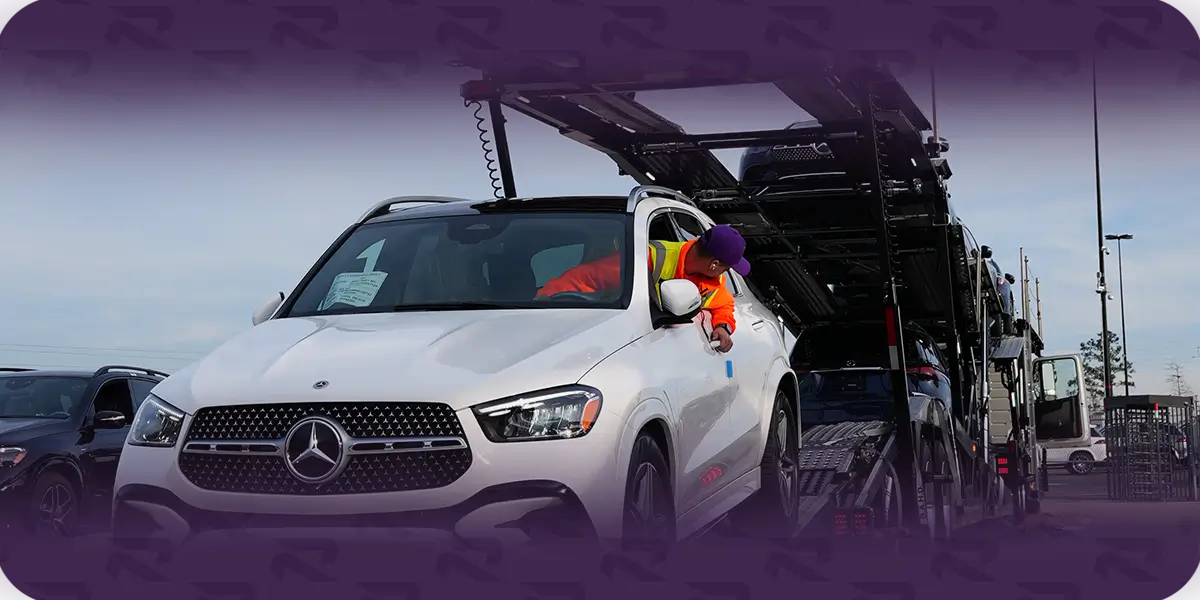 A worker in a hi-vis vest reaching into the driver's window of a white SUV parked beside a loaded open car carrier