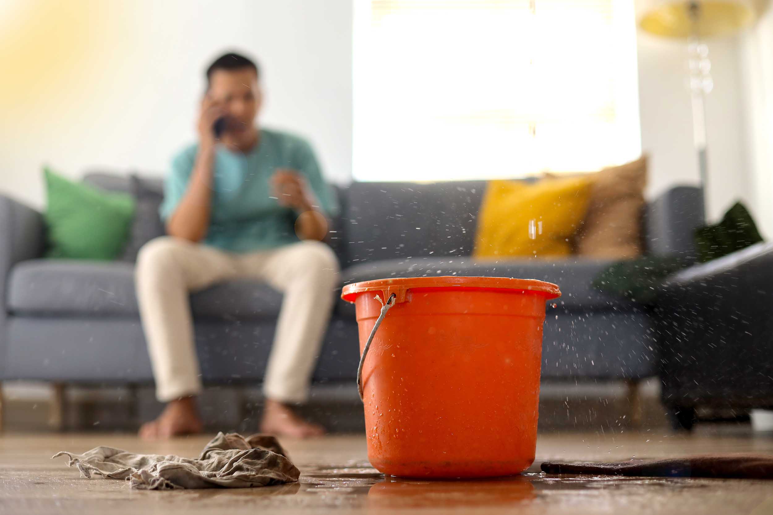 Orange bucket placed on a wet floor with water droplets around, a person sitting on a couch in the background talking on the phone.