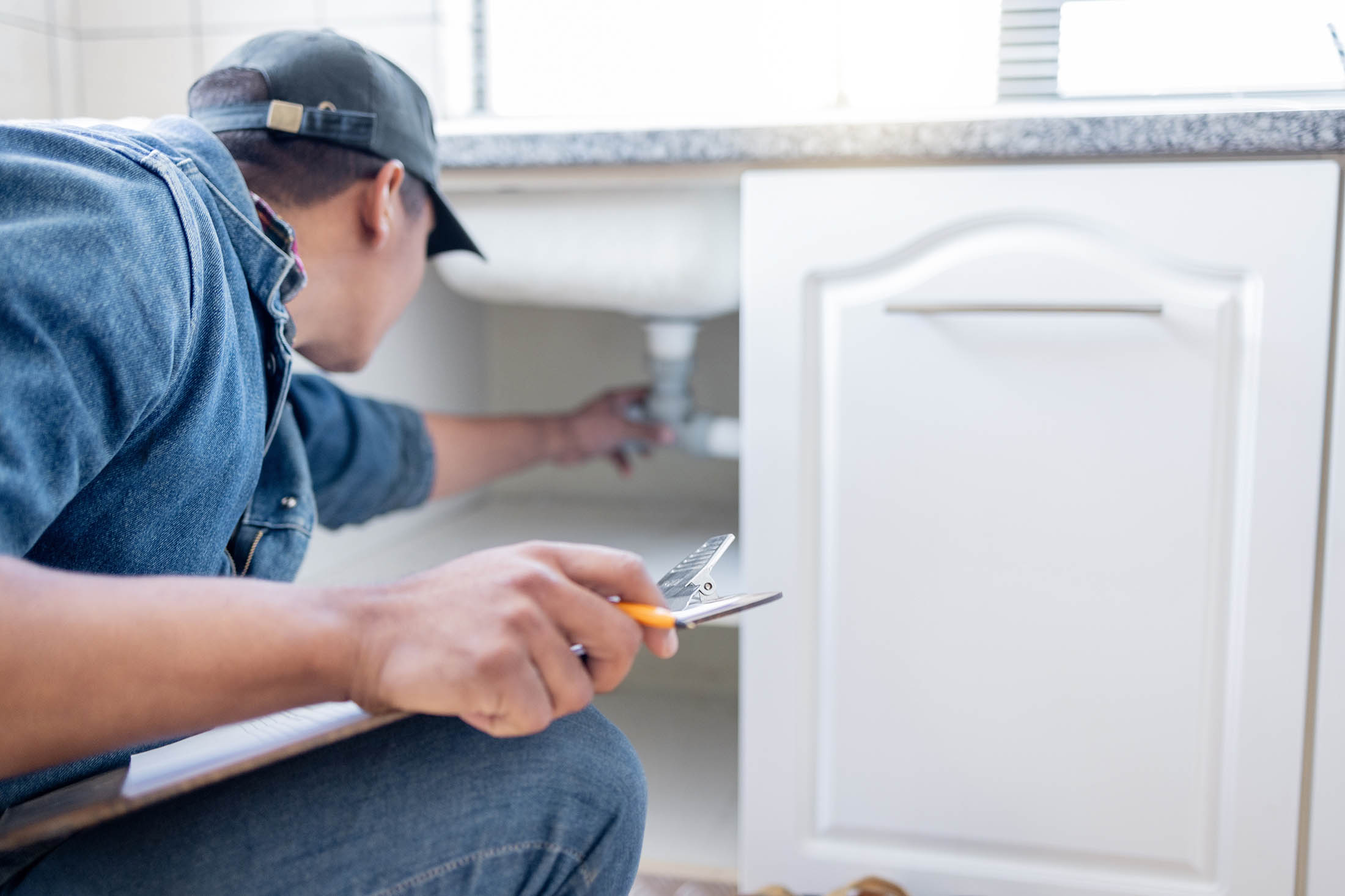 Technician inspecting pipes under a kitchen sink while holding a clipboard.