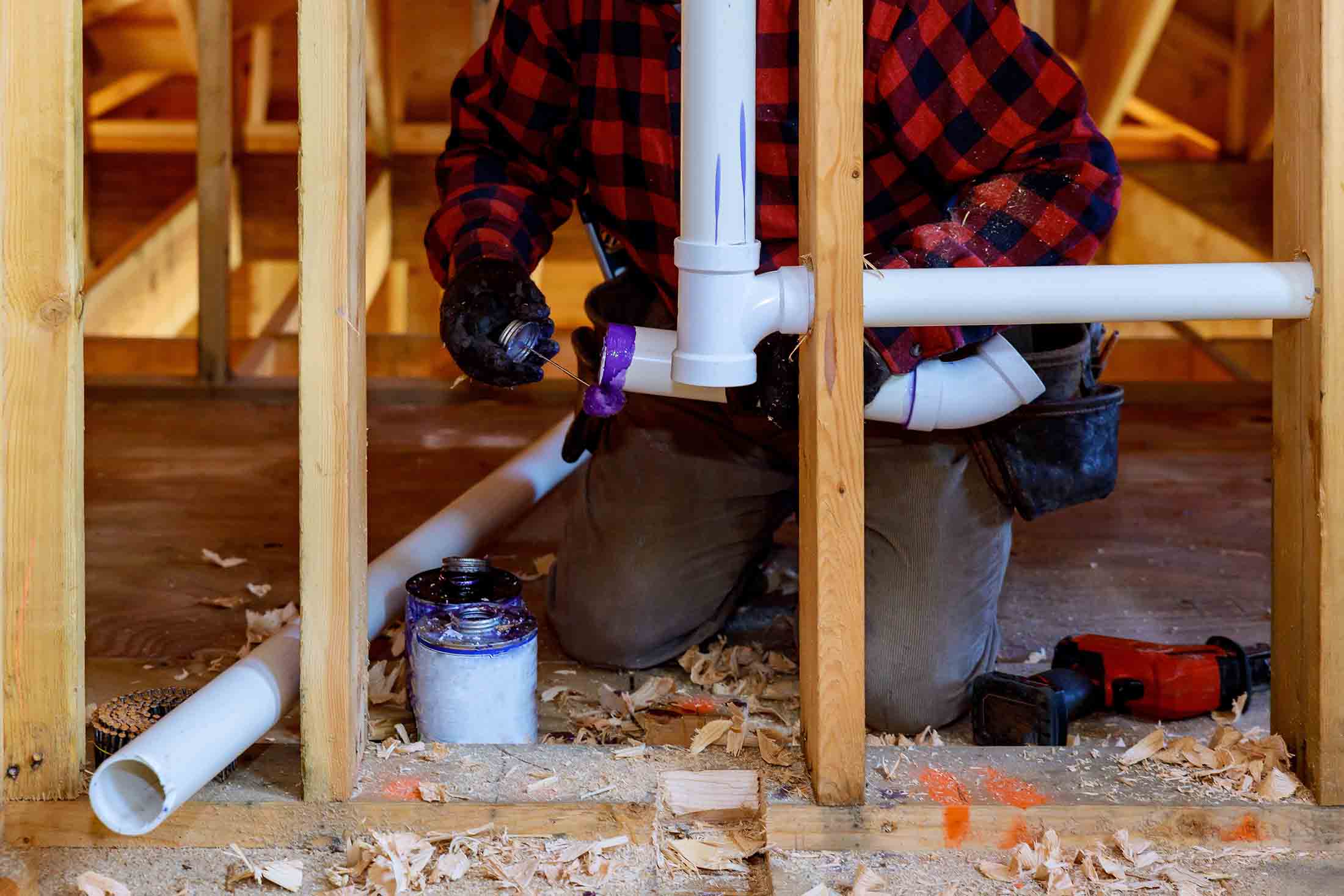 Person in a red and black flannel shirt applying purple primer to white PVC pipes inside wooden framing during construction.