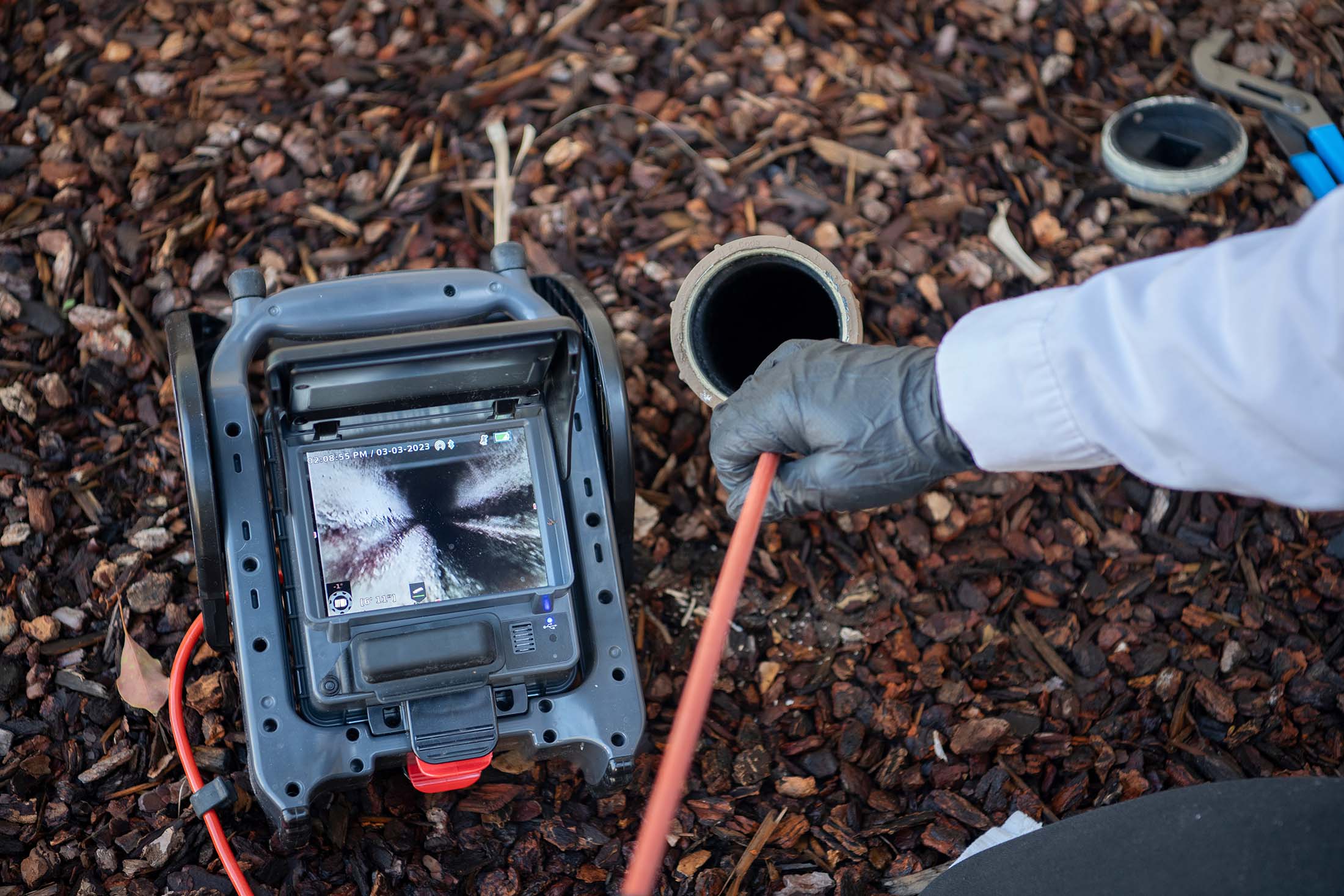 Person wearing a glove inserting an inspection camera cable into an underground pipe with the camera feed visible on a portable monitor.