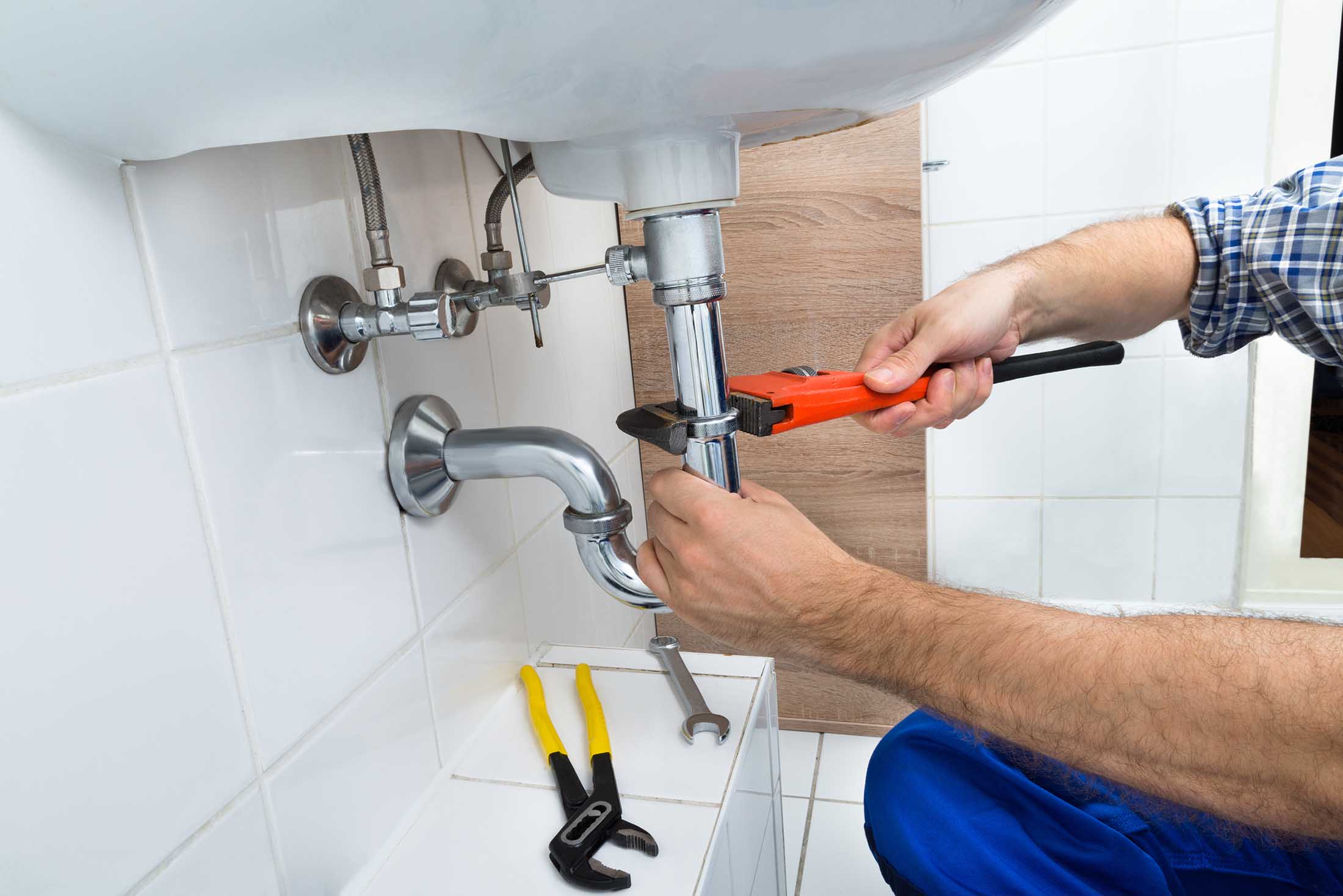 Plumber tightening a chrome pipe under a white sink using a red pipe wrench with other tools nearby.