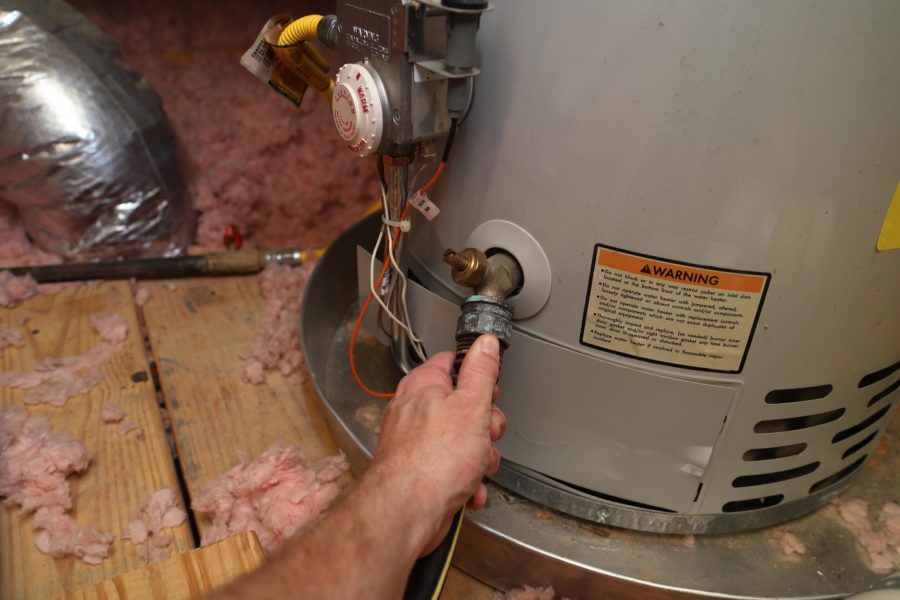 Hand holding a hose connected to the drain valve of a water heater surrounded by pink insulation.