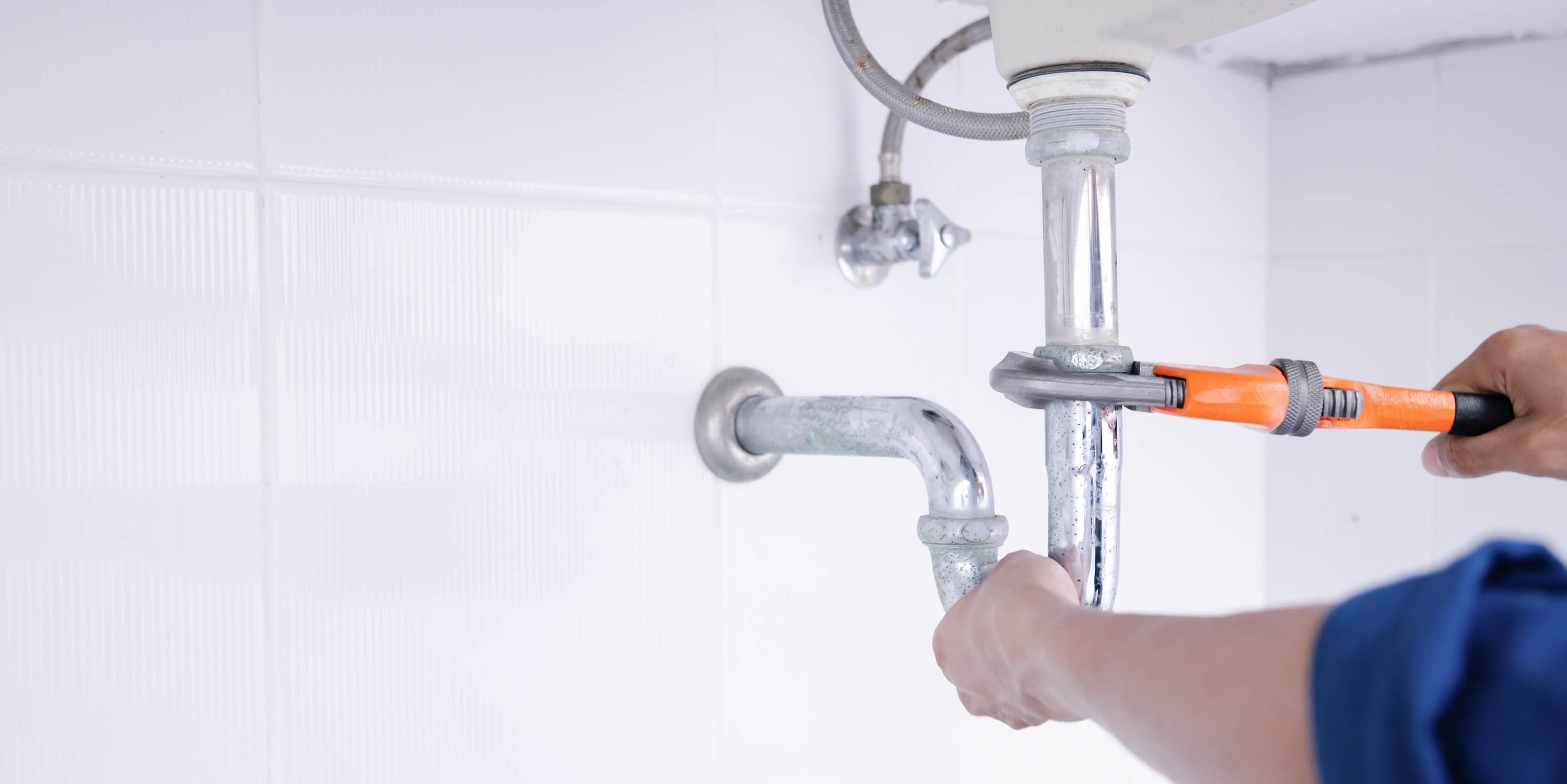 Person using an orange pipe wrench to tighten a chrome sink drain pipe against a white tiled wall.