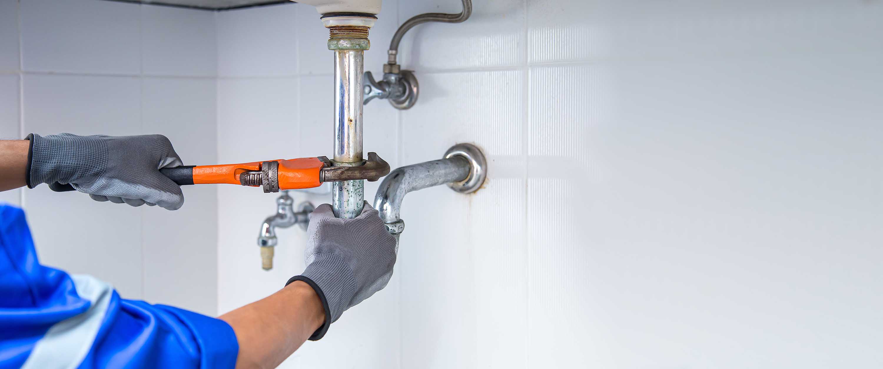 Person wearing gloves using an orange pipe wrench to repair a bathroom drain pipe under a sink.