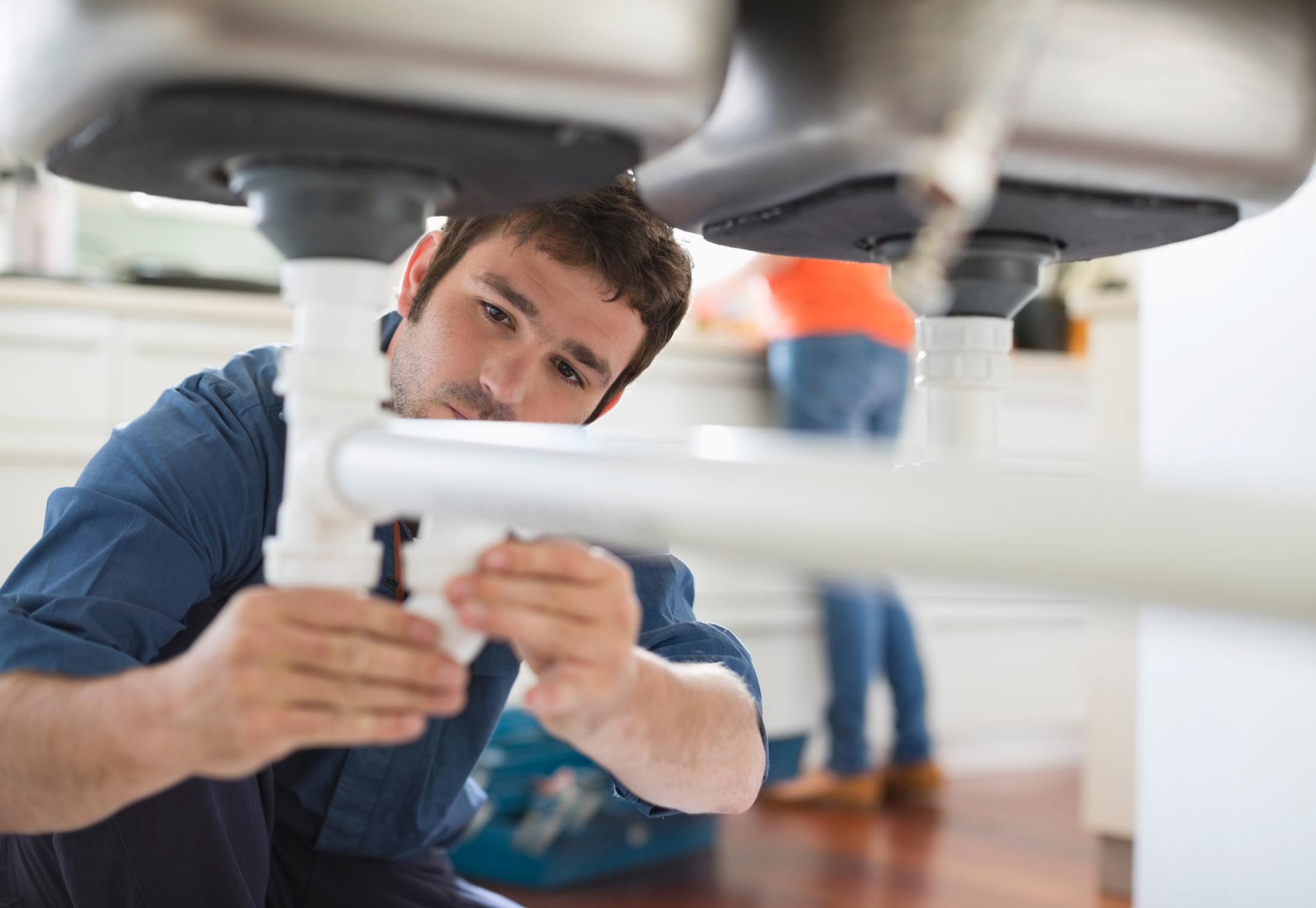 Man in blue shirt fixing white pipes under a kitchen sink with a toolbox in the background.