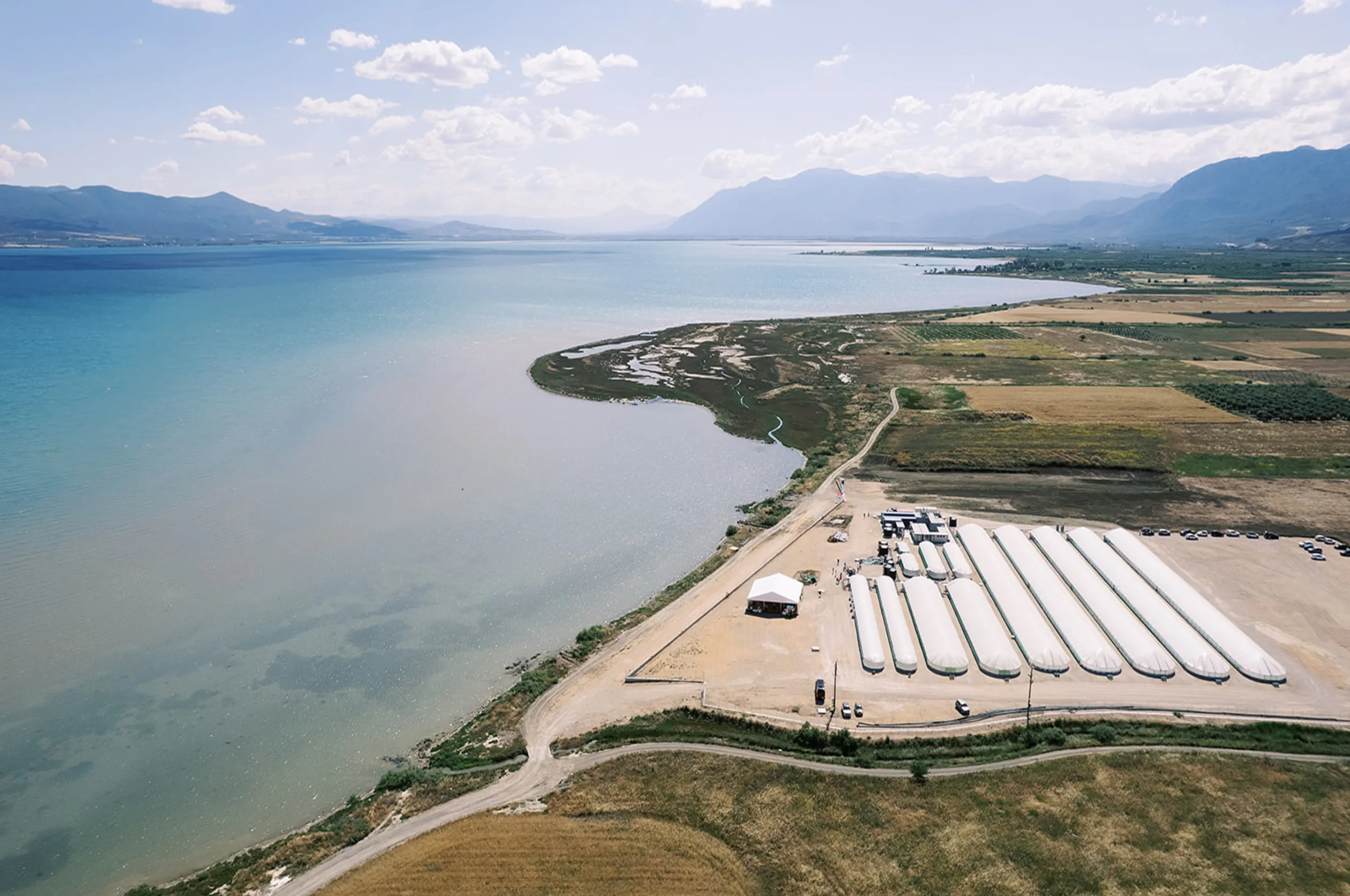 Aerial view of a coastal landscape with a facility featuring long white storage tanks near the shoreline, surrounded by fields and mountains in the background.
