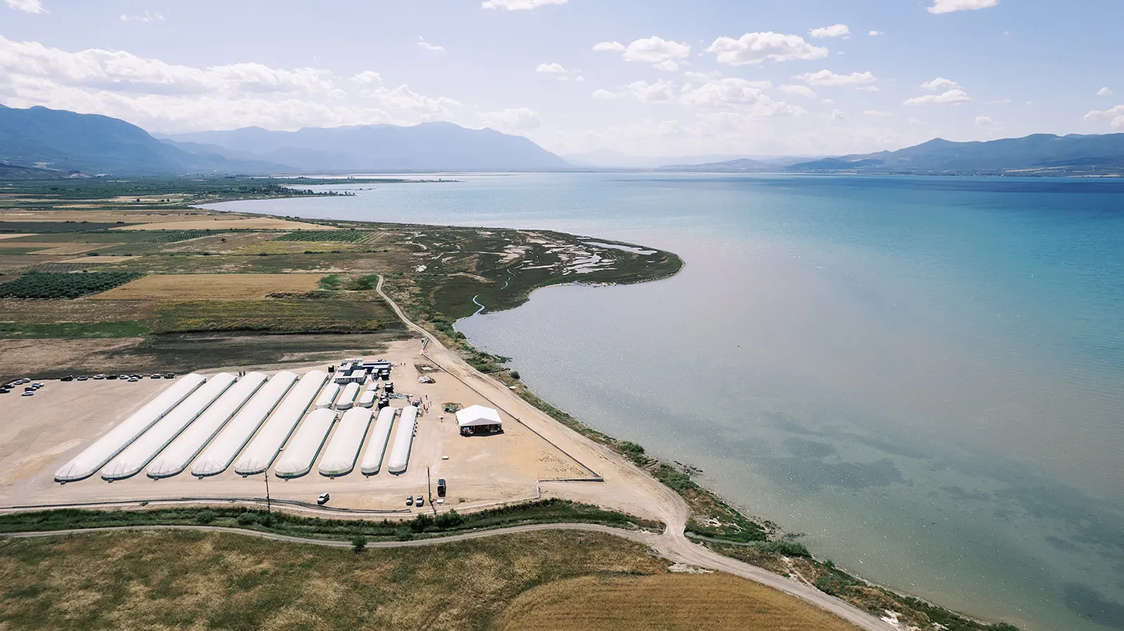 Aerial view of a lakeside facility with long white covered structures, surrounded by agricultural fields and mountains in the background under a partly cloudy sky.