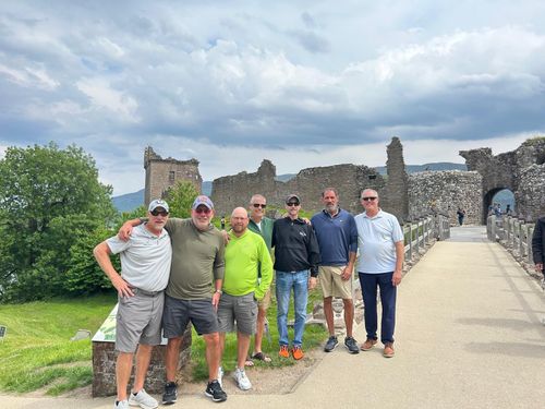 Group of seven men standing together in front of ancient stone castle ruins on a cloudy day.