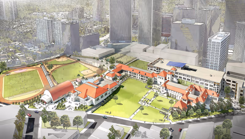 Aerial view of a campus with red-roofed buildings, green sports fields, and tall city skyscrapers in the background.