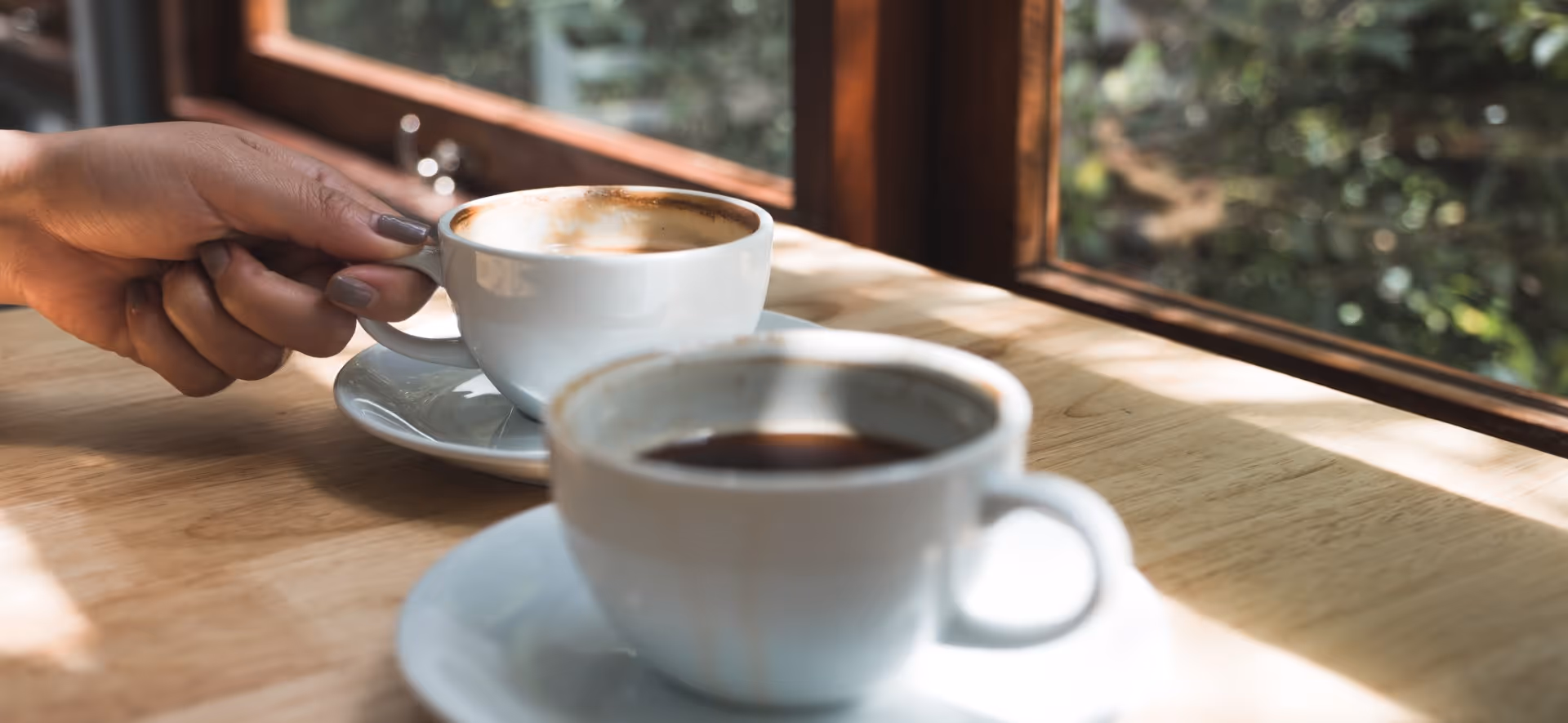Two white coffee cups on wooden table near window with soft sunlight