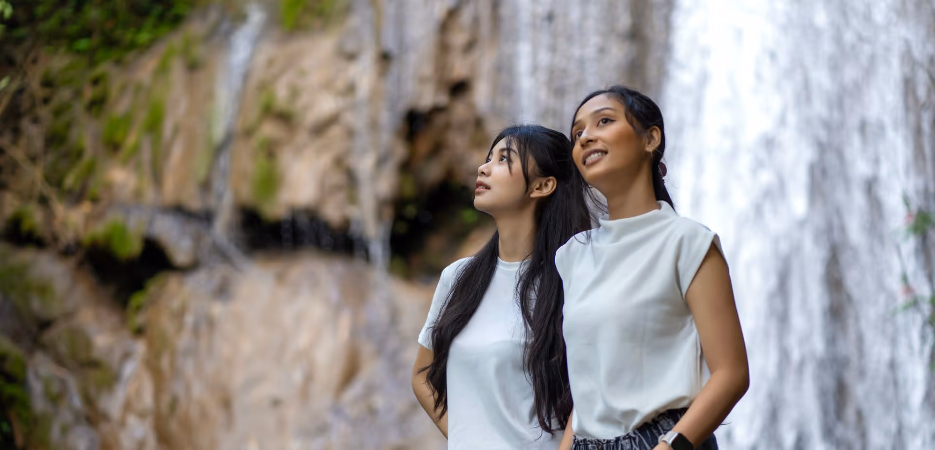 Two women in white tops standing together near rocky waterfall backdrop