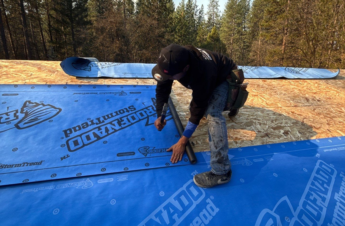 Worker installing blue waterproof roofing underlayment on wooden roof decking outdoors.