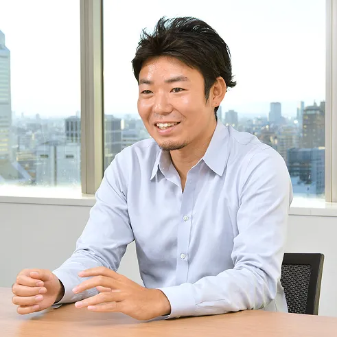 Man in a light blue shirt smiling and gesturing while sitting at a table in an office with city buildings visible through large windows behind him.