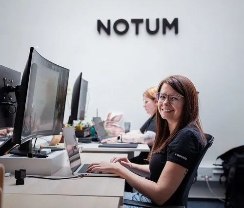 Smiling woman with glasses typing on a laptop at an office desk with 'NOTUM' sign on the wall behind her.