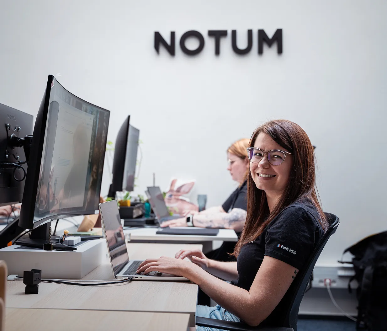Smiling woman with glasses typing on a laptop at an office desk with 'NOTUM' sign on the wall behind her.