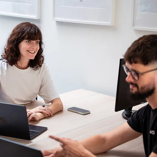 Two colleagues collaborate at a desk with laptops, one woman smiling and listening while a man gestures.