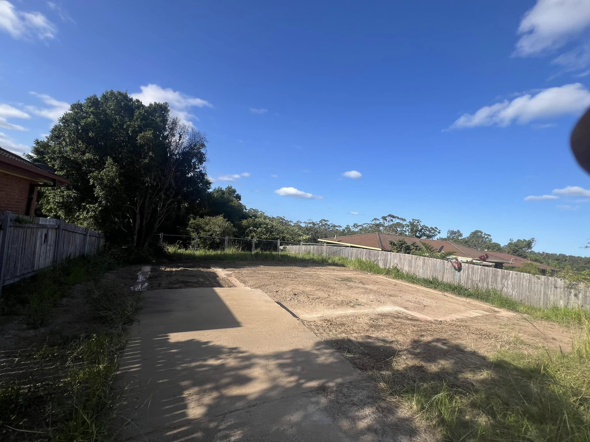 Cleared backyard with fence, trees, and neighboring rooftops under a blue sky with scattered clouds.