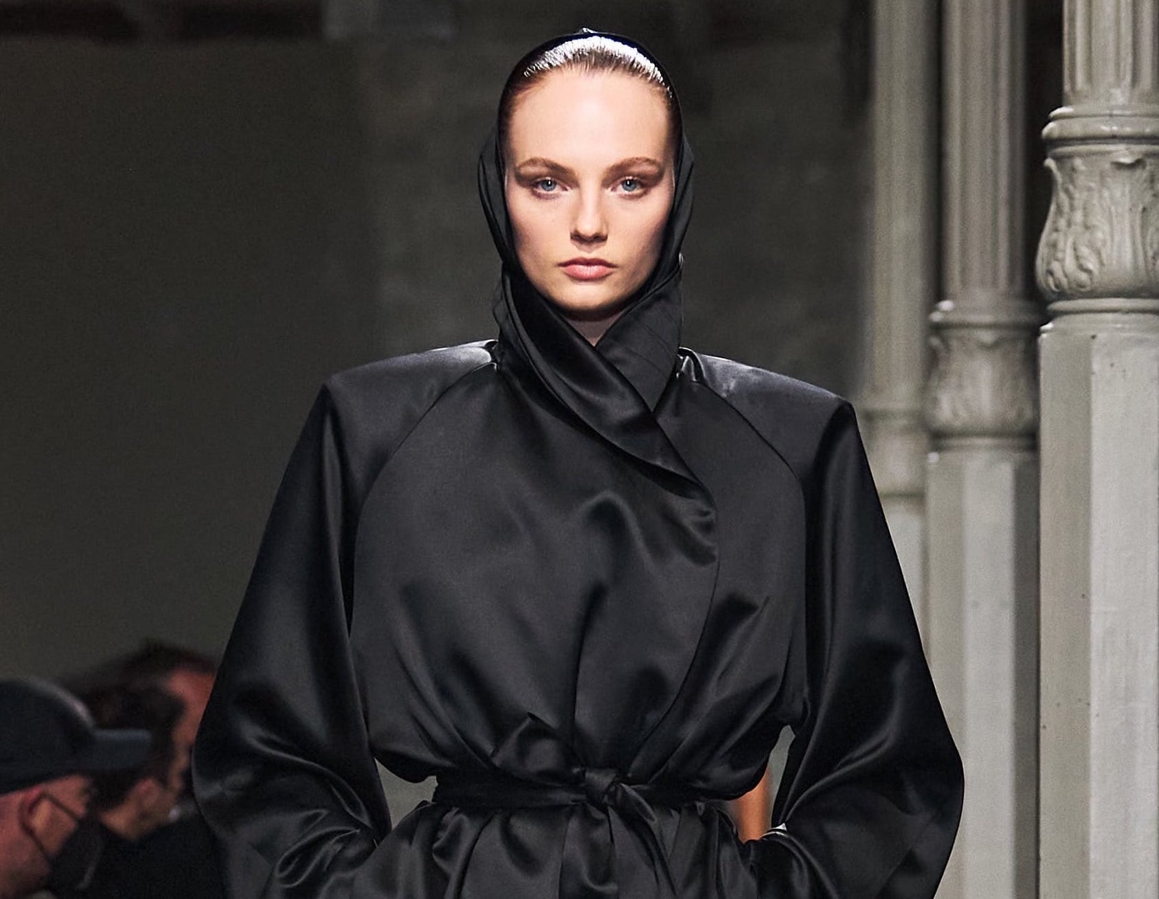 Fashion model wearing a sleek black satin robe with a high collar and tied waist on a runway.