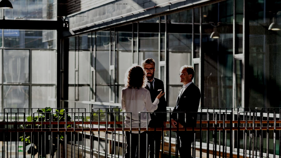 Three business professionals having a discussion in a modern office atrium with large glass windows.