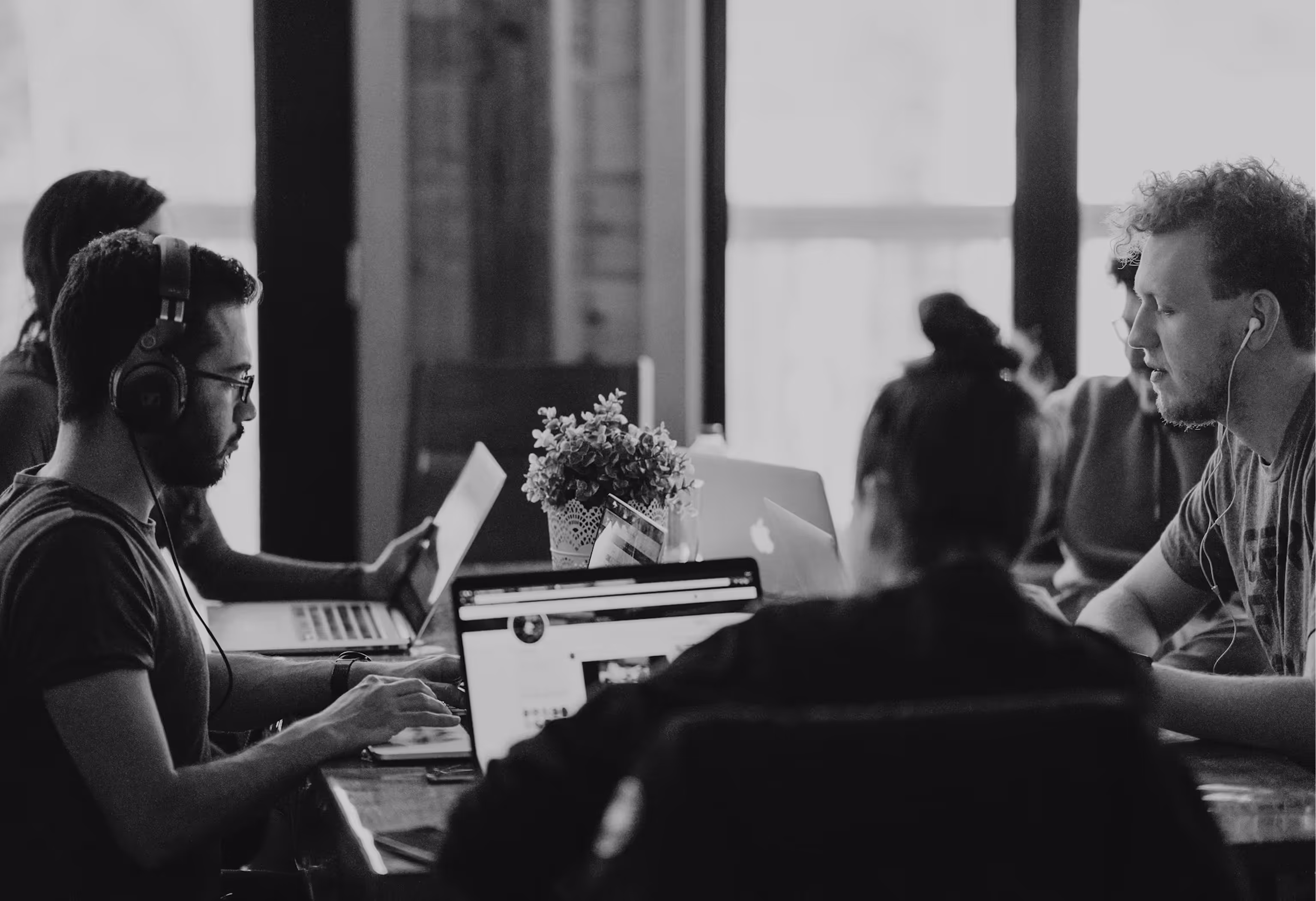 Group of young professionals working on laptops around a table in a modern office setting.