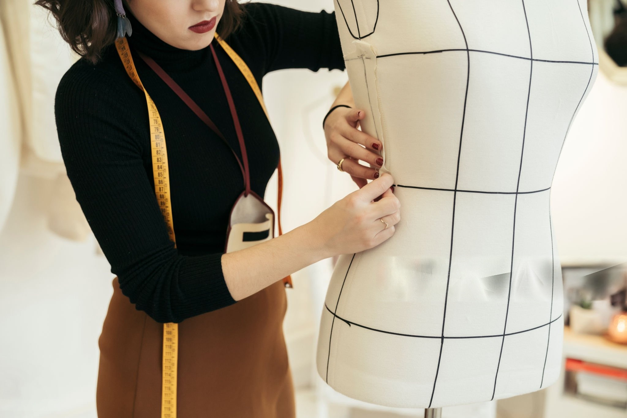 Woman adjusting fabric on a dress form marked with black grid lines, wearing a black top and brown skirt with a yellow measuring tape around her neck.
