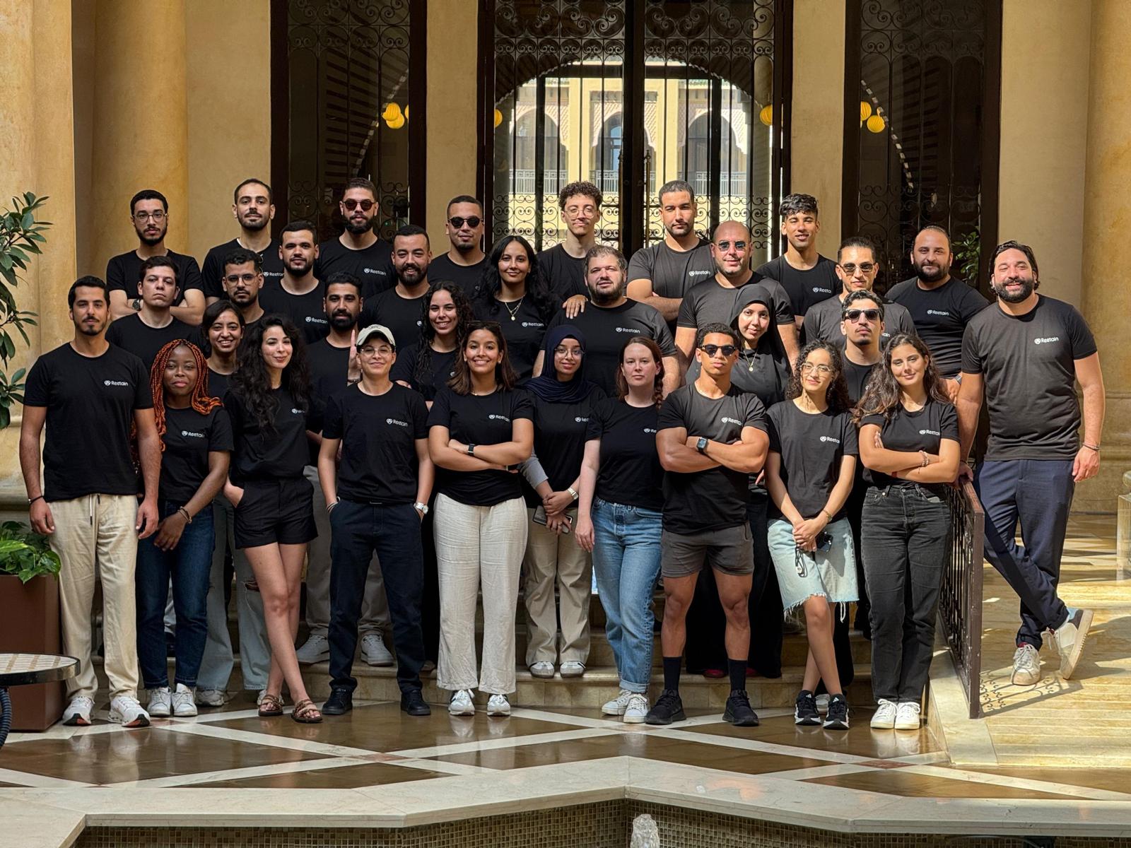 Group of approximately 35 people wearing black Retoin t-shirts posing indoors in front of an ornate gated entrance with columns.