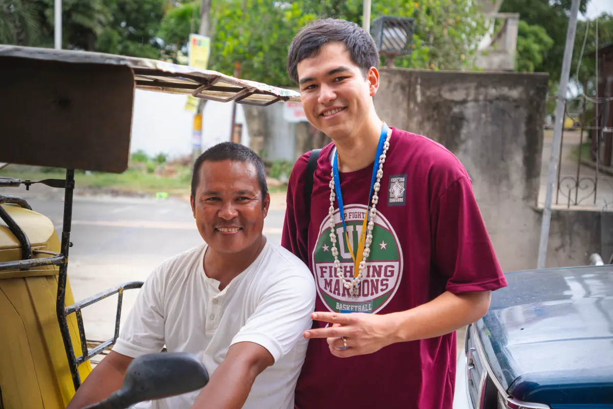 Smiling man in white shirt sitting in a vehicle with a younger man in a maroon shirt standing beside him making a peace sign.