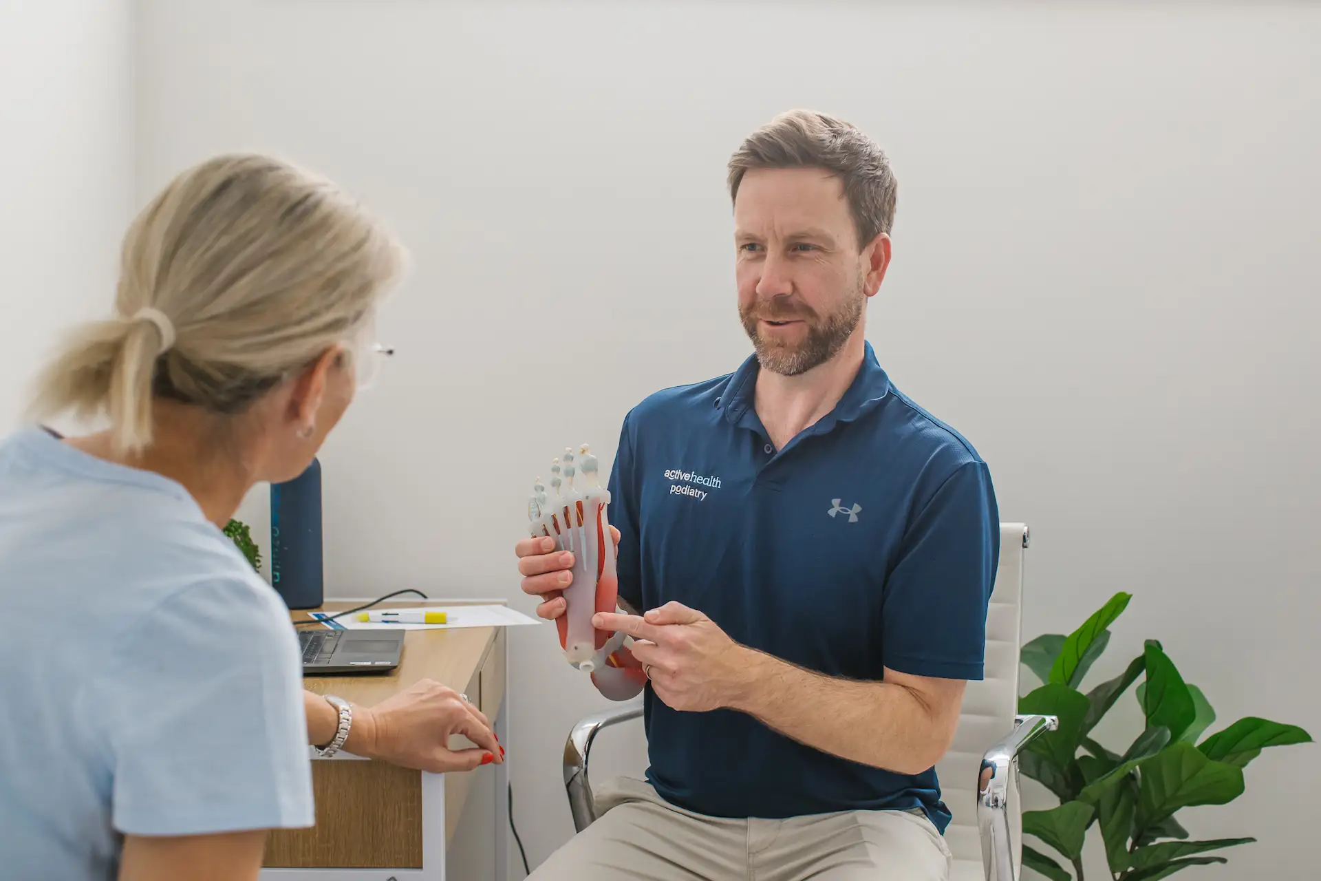 Male podiatrist in blue polo shirt holding and explaining a foot anatomy model to a female patient in an office.