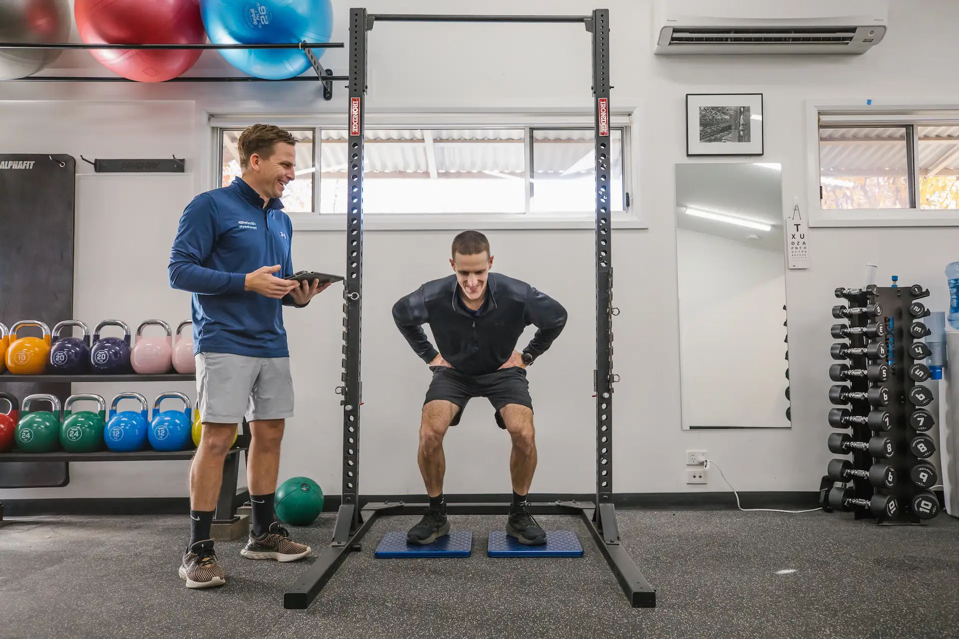 A man performing a squat exercise on blue pads inside a gym while a trainer holds a tablet and observes him.