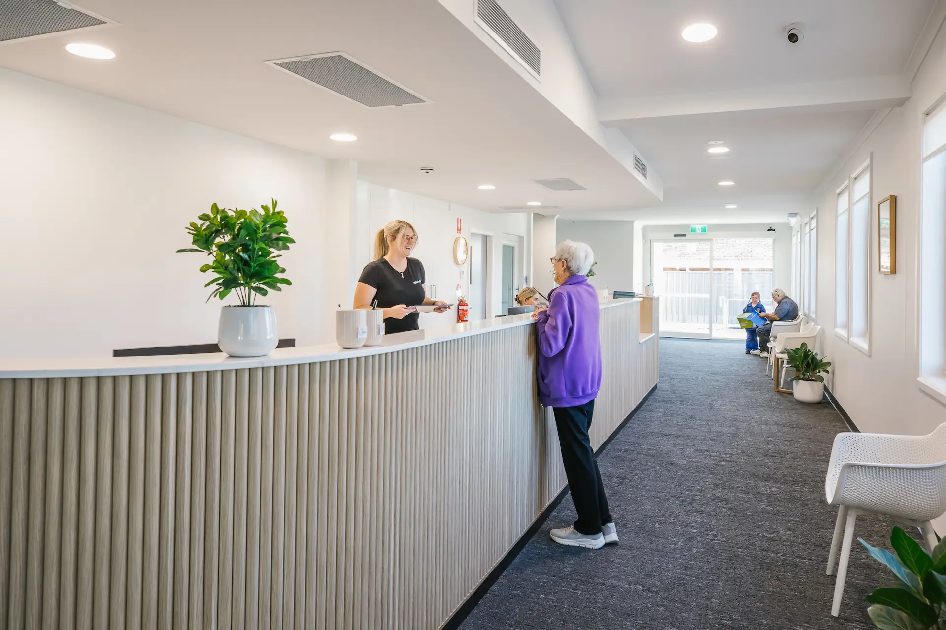 Modern reception area with a smiling receptionist assisting an elderly woman, seating area with two people talking in the background.
