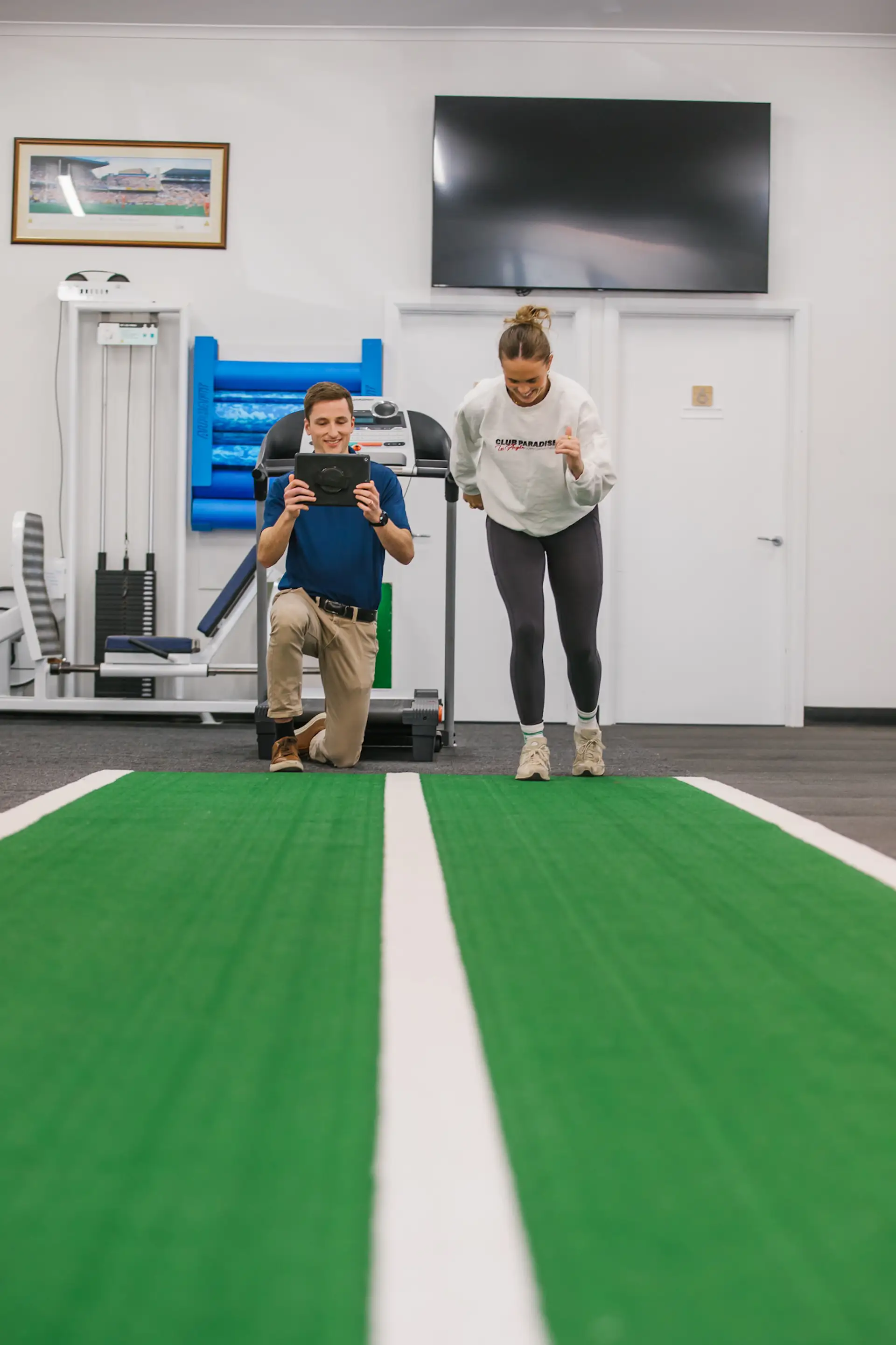 A man kneeling with a tablet records a woman jogging indoors on a green track in a fitness facility.