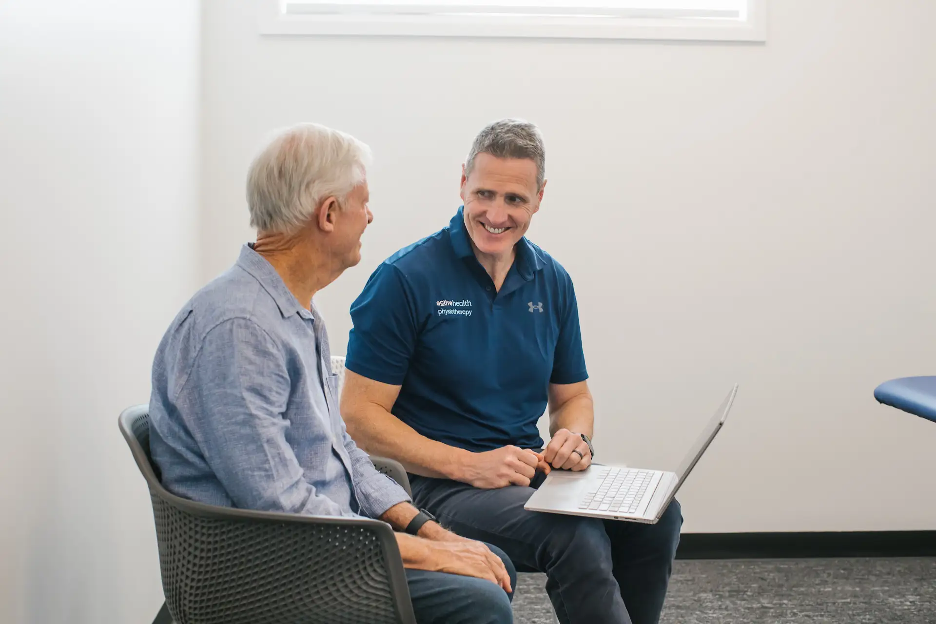 Physiotherapist smiling and showing something on a laptop while consulting an elderly man in an office.