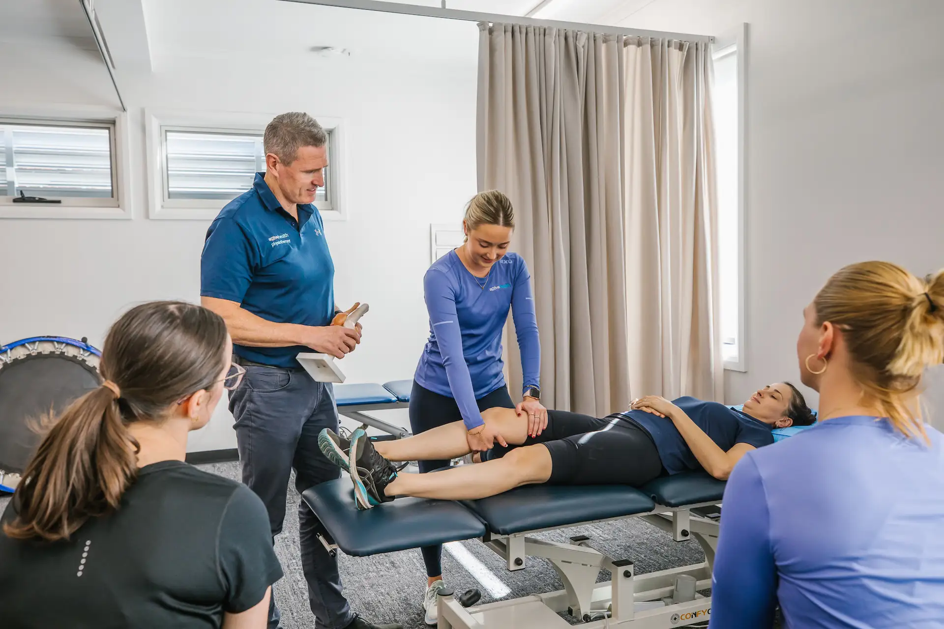 Physiotherapist demonstrating knee exercise on a patient lying on a treatment table while two others observe in a clinical room.