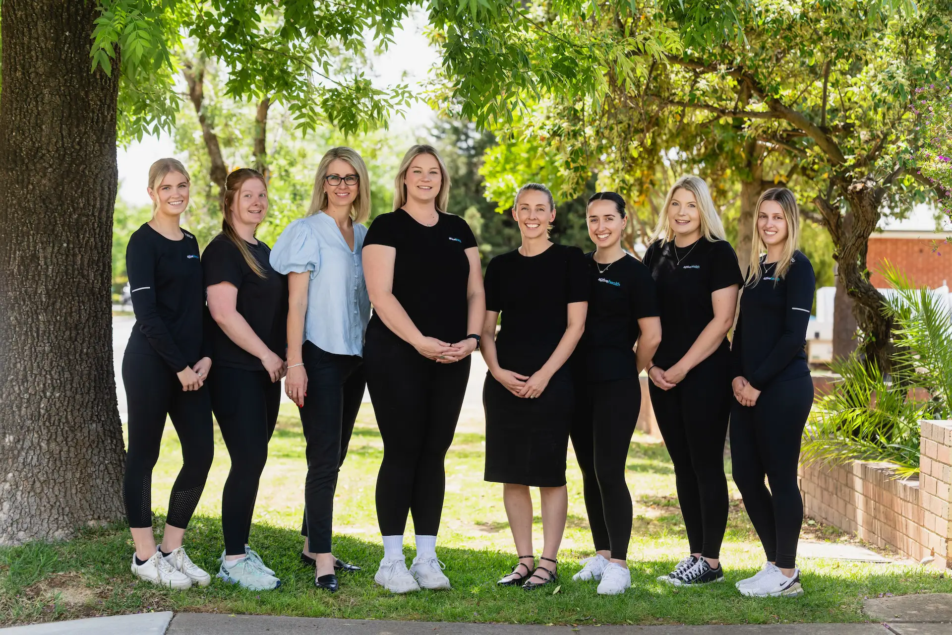 Group of eight women standing outdoors on grass under trees, smiling at the camera.